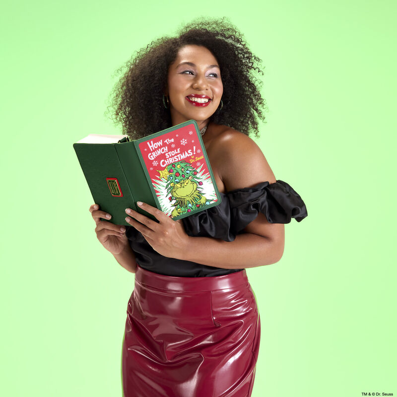 Woman wearing a black off-the-shoulder top and red leather skirt, holding the Loungefly How the Grinch Stole Christmas crossbody bag as though she&rsquo;s reading a book. She stands against a green background. 